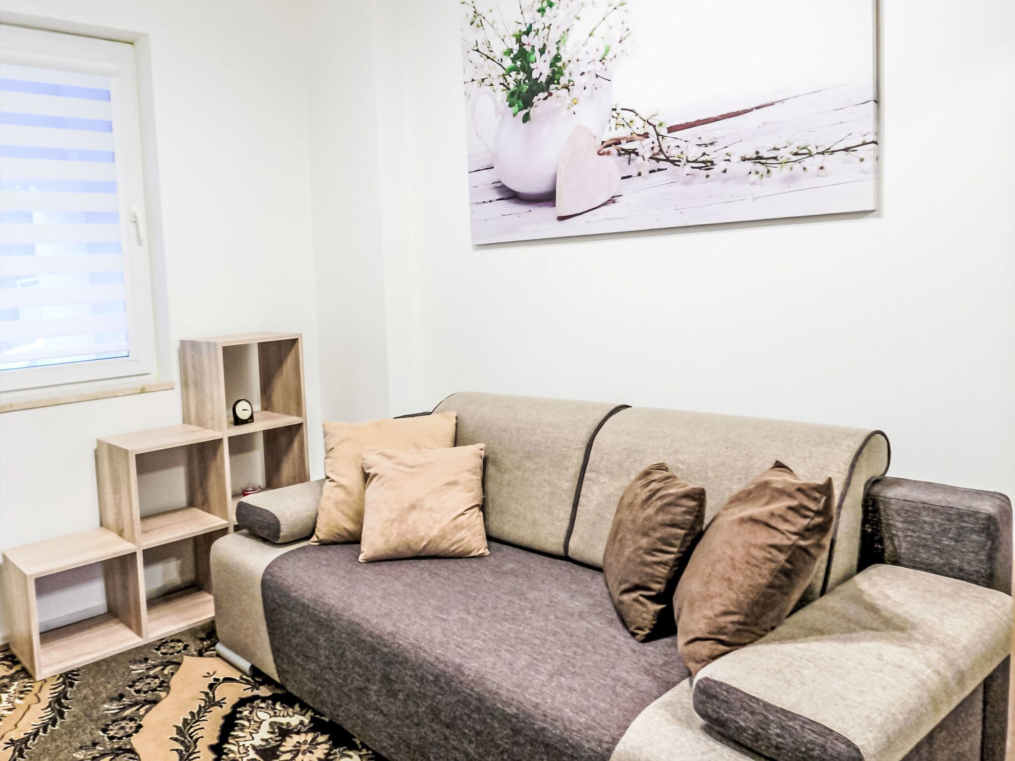 A modern living room with a grey sofa, beige cushions, a small wooden side table, a wall-mounted shelf, and a framed picture of flowers.