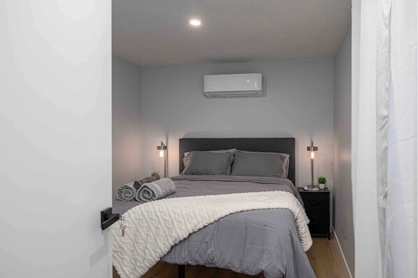 A bedroom with a bed, two bedside tables, a wall-mounted air conditioner, and a ceiling light.