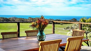 Une table en bois sur laquelle est posé un vase de fleurs, quatre chaises en bois, et une vue sur l’océan et les collines.