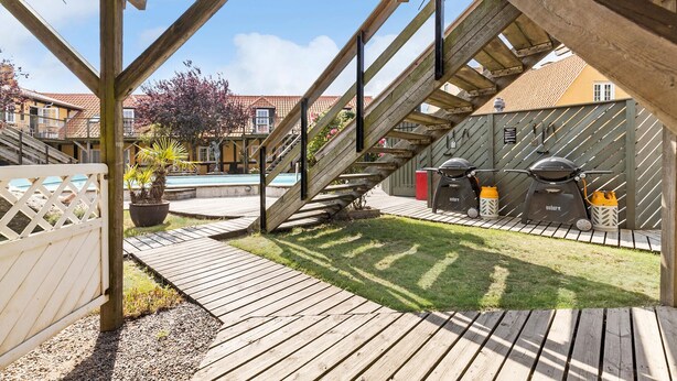 Una terraza de madera con zona de barbacoa, piscina y escalera que lleva a un segundo piso.