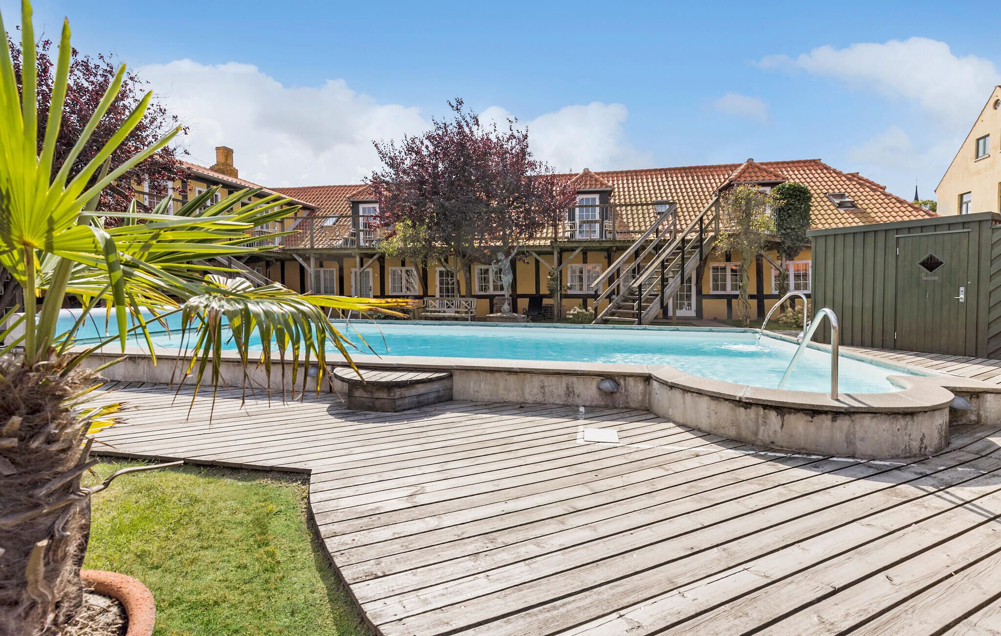 A wooden deck with a swimming pool, a palm tree, and a building with a tiled roof in the background.