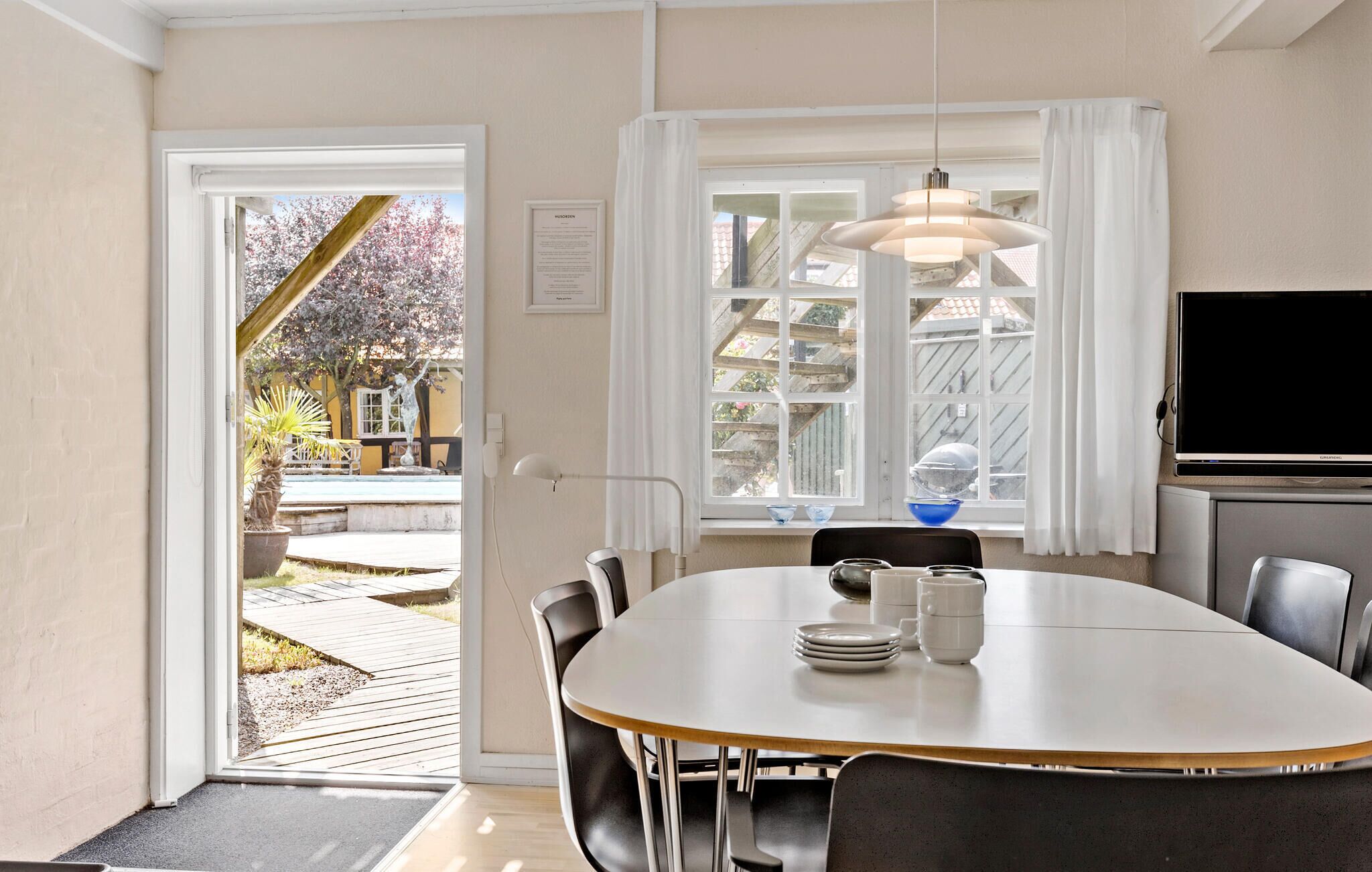 A dining area with a large table, chairs, a hanging light fixture, and a view of a garden through a window.
