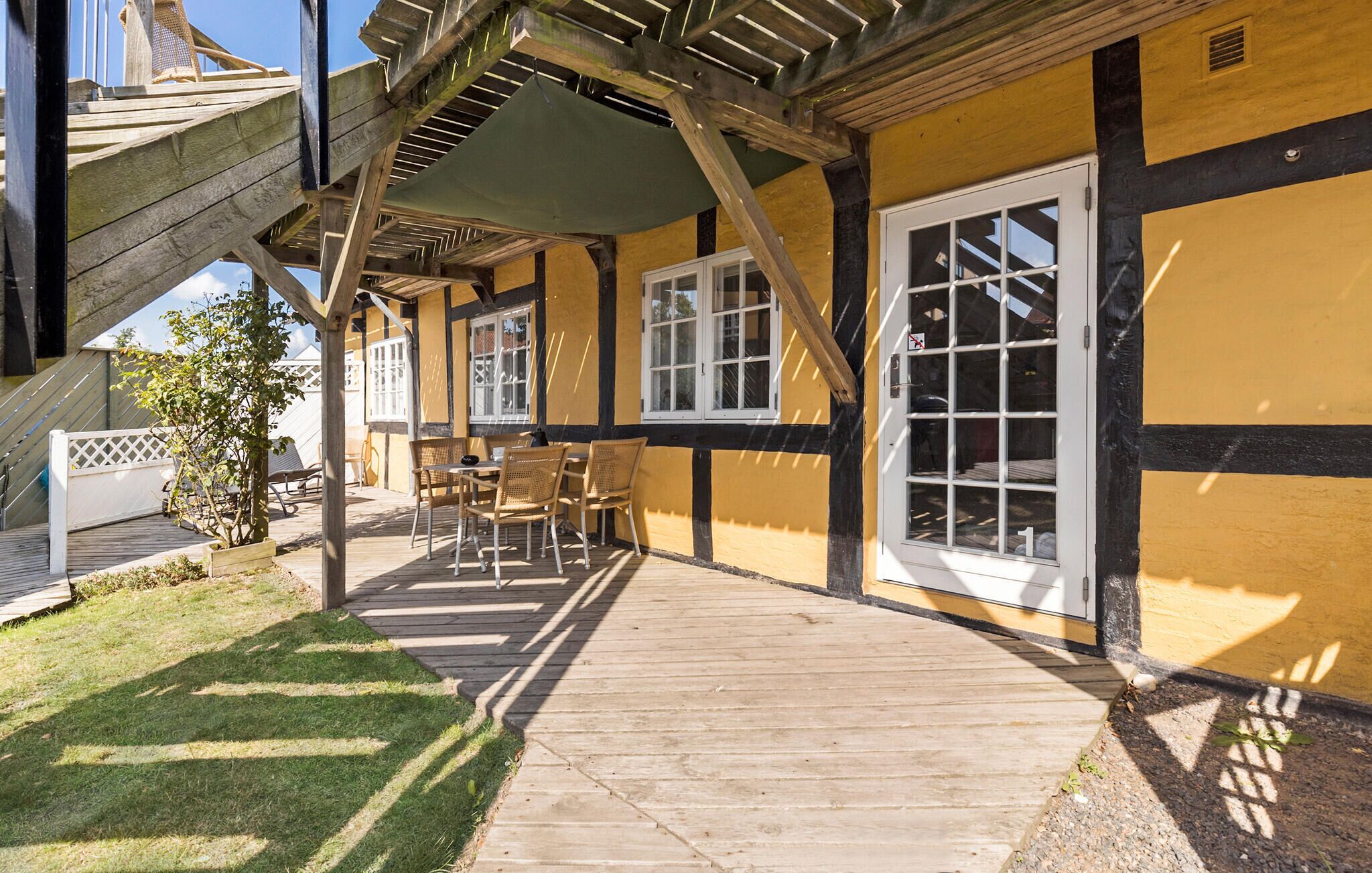 Ein überdachter Patio-Bereich mit Holzdeck, Sitzgelegenheiten im Freien und Blick auf ein gelbes Gebäude mit schwarzer Verzierung und weißen Fenstern.