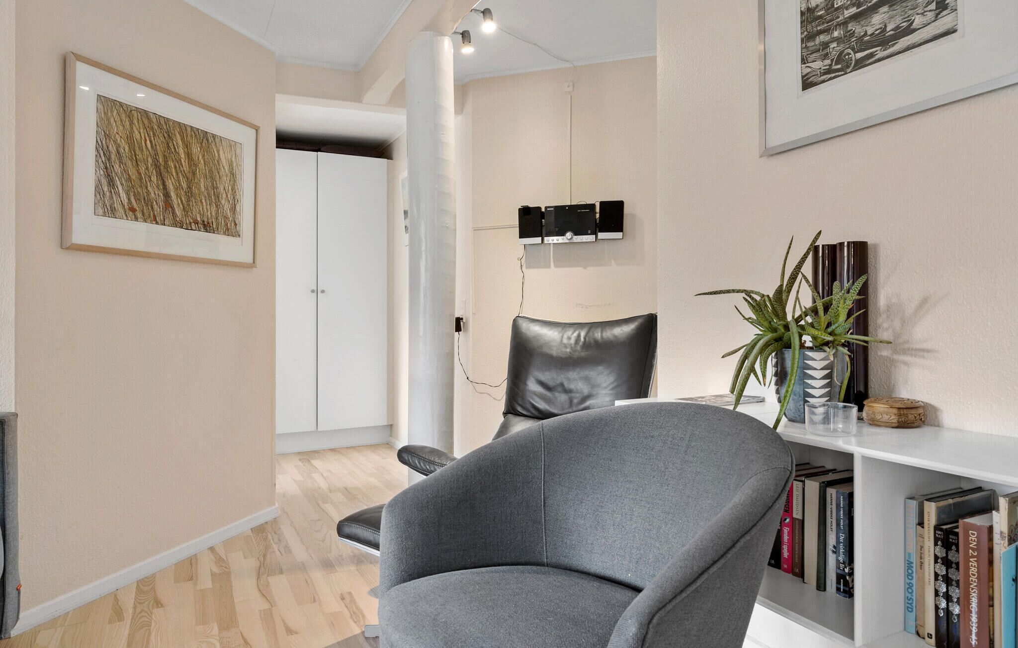A modern living room with a grey armchair, a bookshelf with books, a framed artwork, a white wardrobe, and a mounted television.