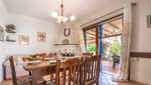 A dining area with a wooden table set for four, surrounded by chairs with patterned cushions. A chandelier hangs above the table, and a window with a view of greenery is visible. - Le palme by Interhome (Villasimius)