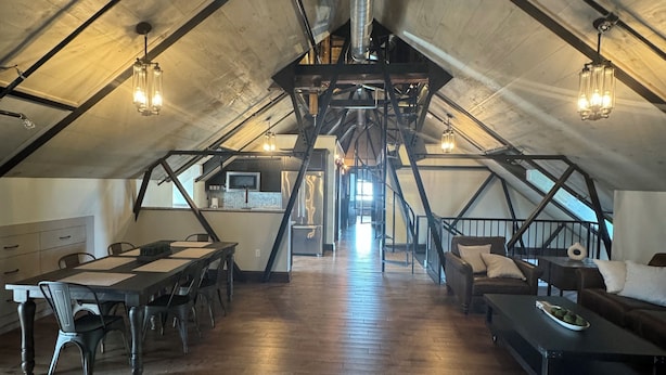 Un espace mansardé avec une haute voûte, des poutres apparentes et un escalier central. On y trouve une longue table à manger avec des chaises, ainsi qu’un coin salon avec un canapé et une table basse.