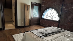 Une chambre avec un plancher en bois, un lit recouvert d’une courtepointe, une porte avec un panneau de verre et un mur en briques.