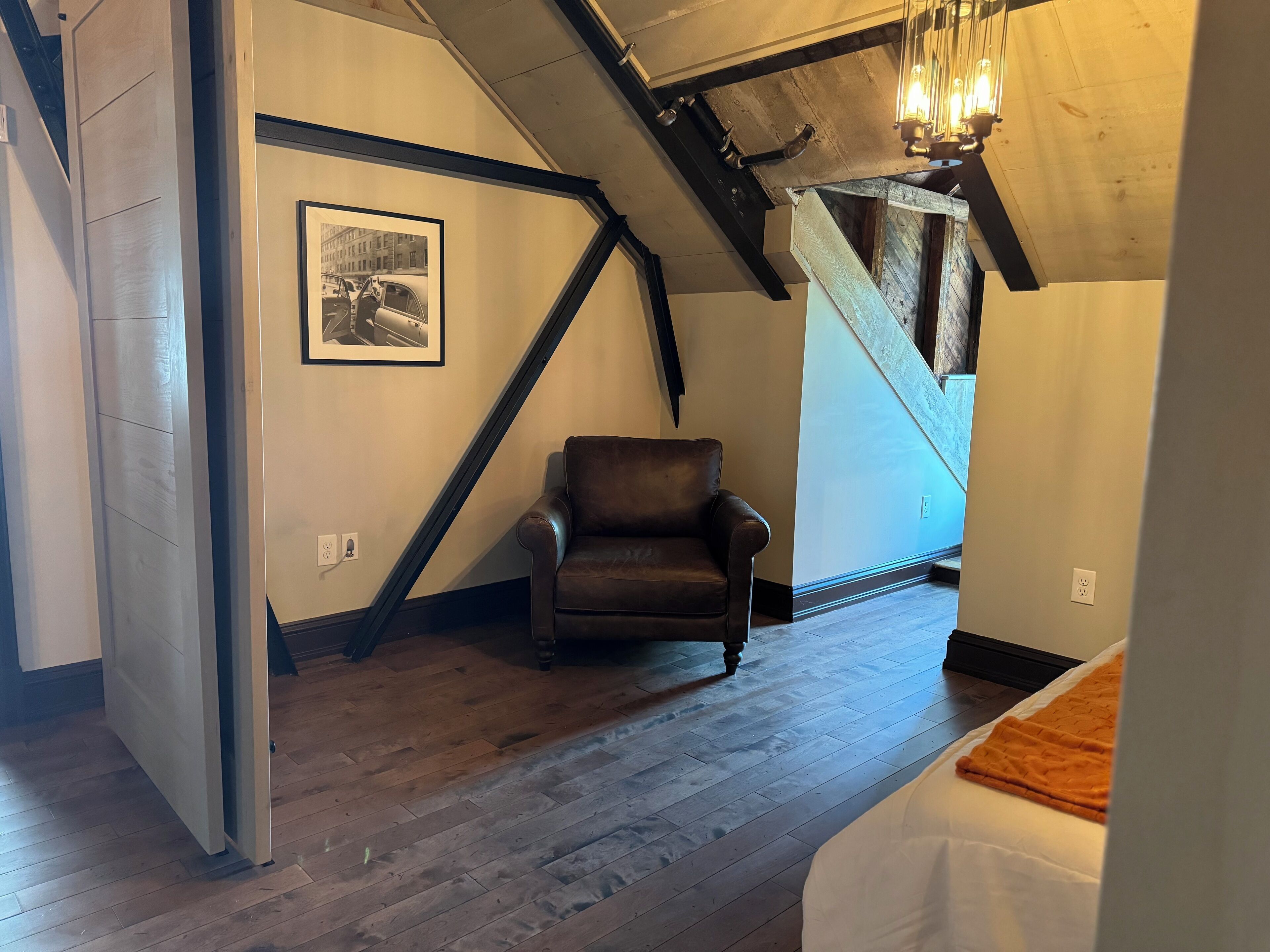 A room with a wooden floor, a brown leather chair, a framed picture on the wall, and a staircase with exposed beams.