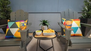 Zwei Adirondack-Stühle mit bunten Kissen, ein kleiner Tisch mit einer Schüssel Chips und Salsa sowie ein Blumentopf.