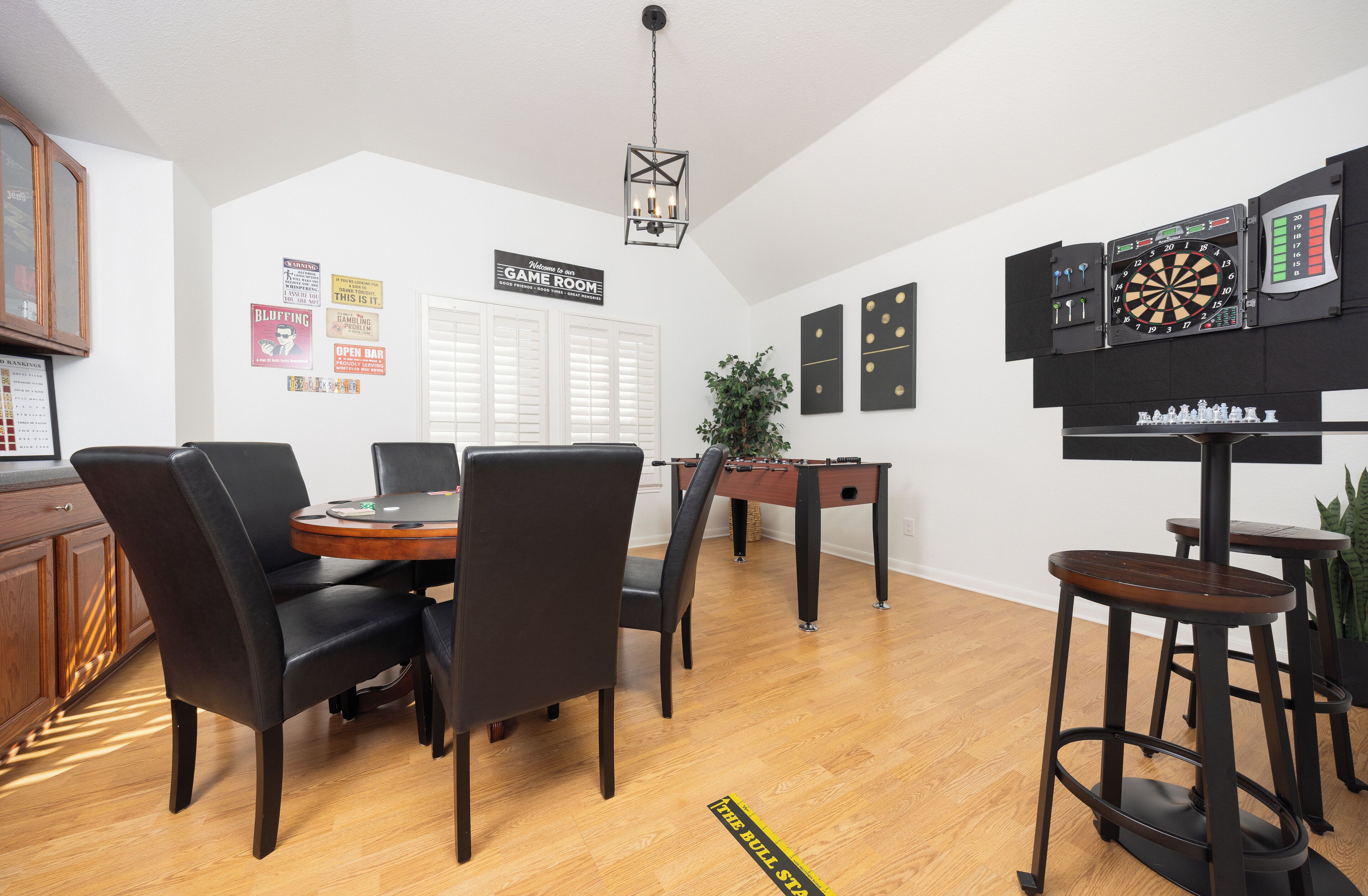 A room with a wooden dining table, black chairs, a foosball table, a dartboard, and a chessboard.