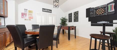 A room with a wooden dining table, black chairs, a foosball table, a dartboard, and a chessboard.