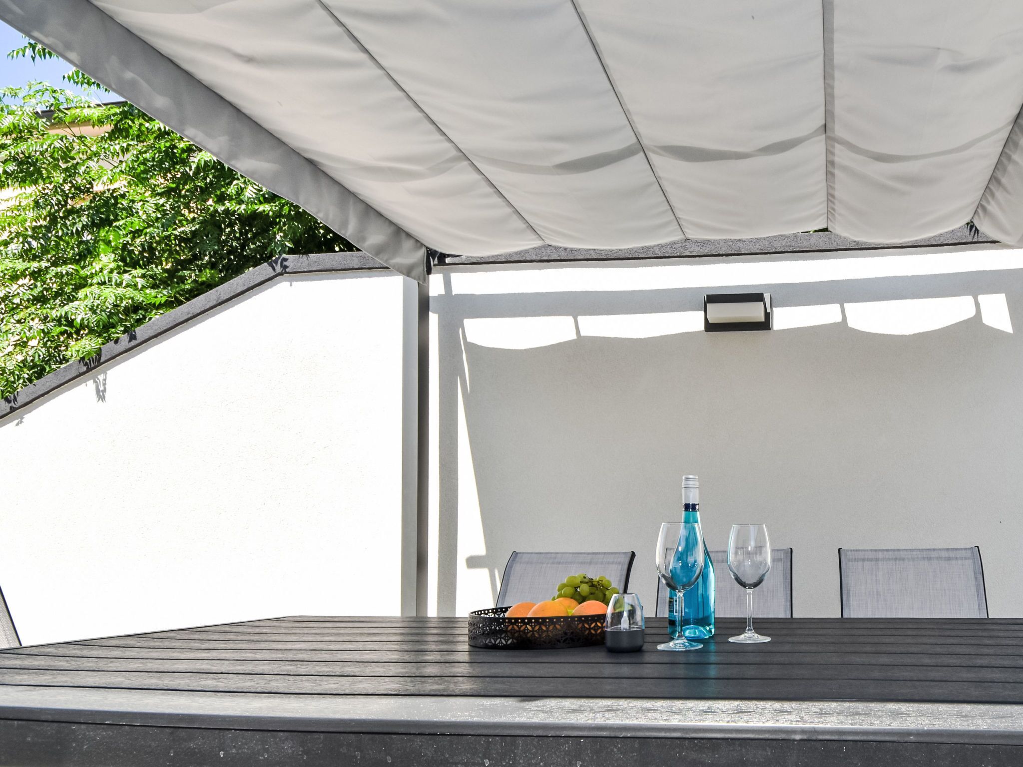 A covered outdoor seating area with a table, chairs, a basket of fruit, and a bottle of water.