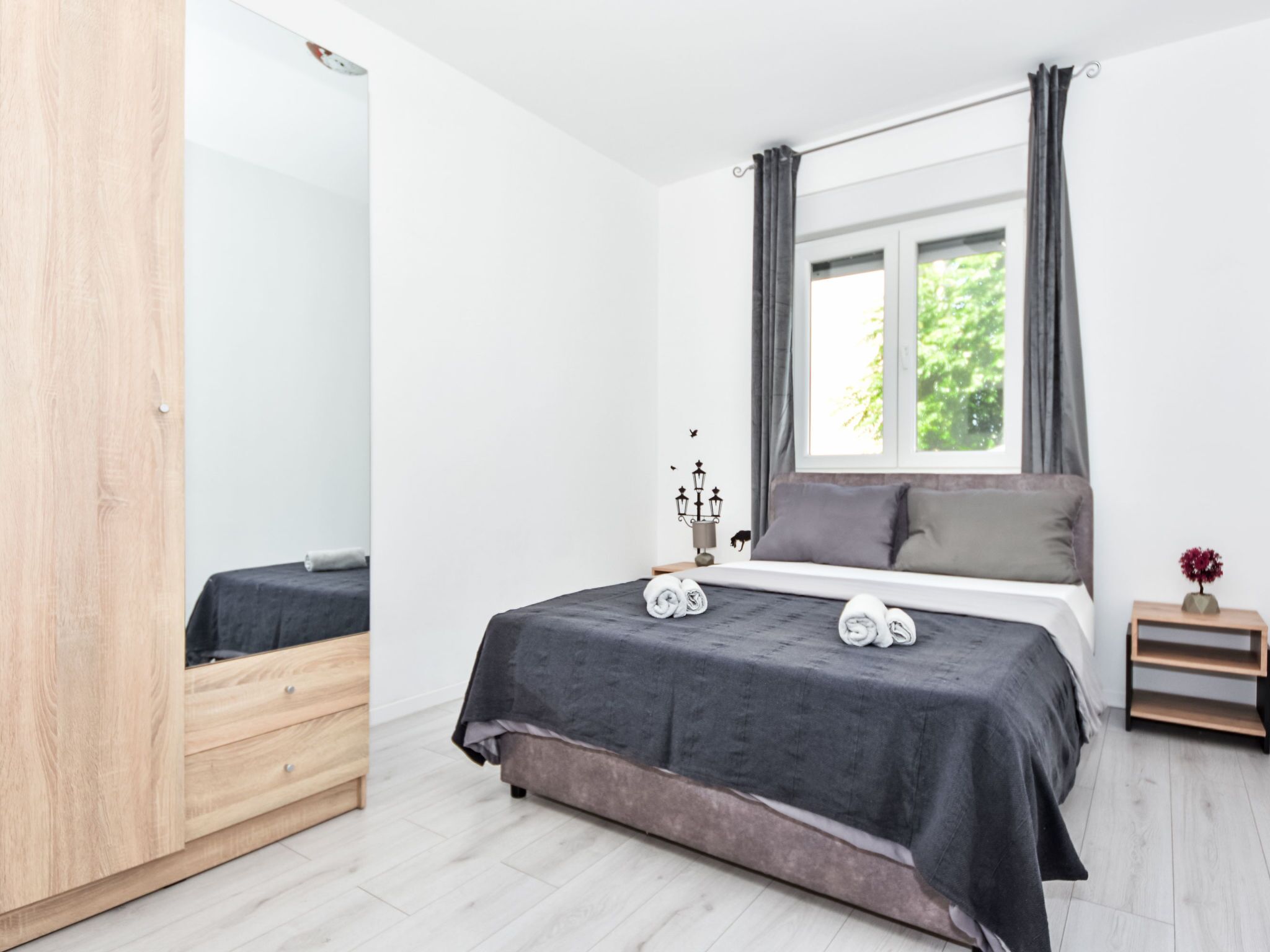 A bedroom with a bed, a nightstand, a mirror, and a window with curtains.