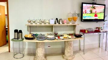 Disposição de buffet com diversos alimentos e bebidas sobre uma mesa de mármore, um canto para café e uma televisão exibindo um evento esportivo.