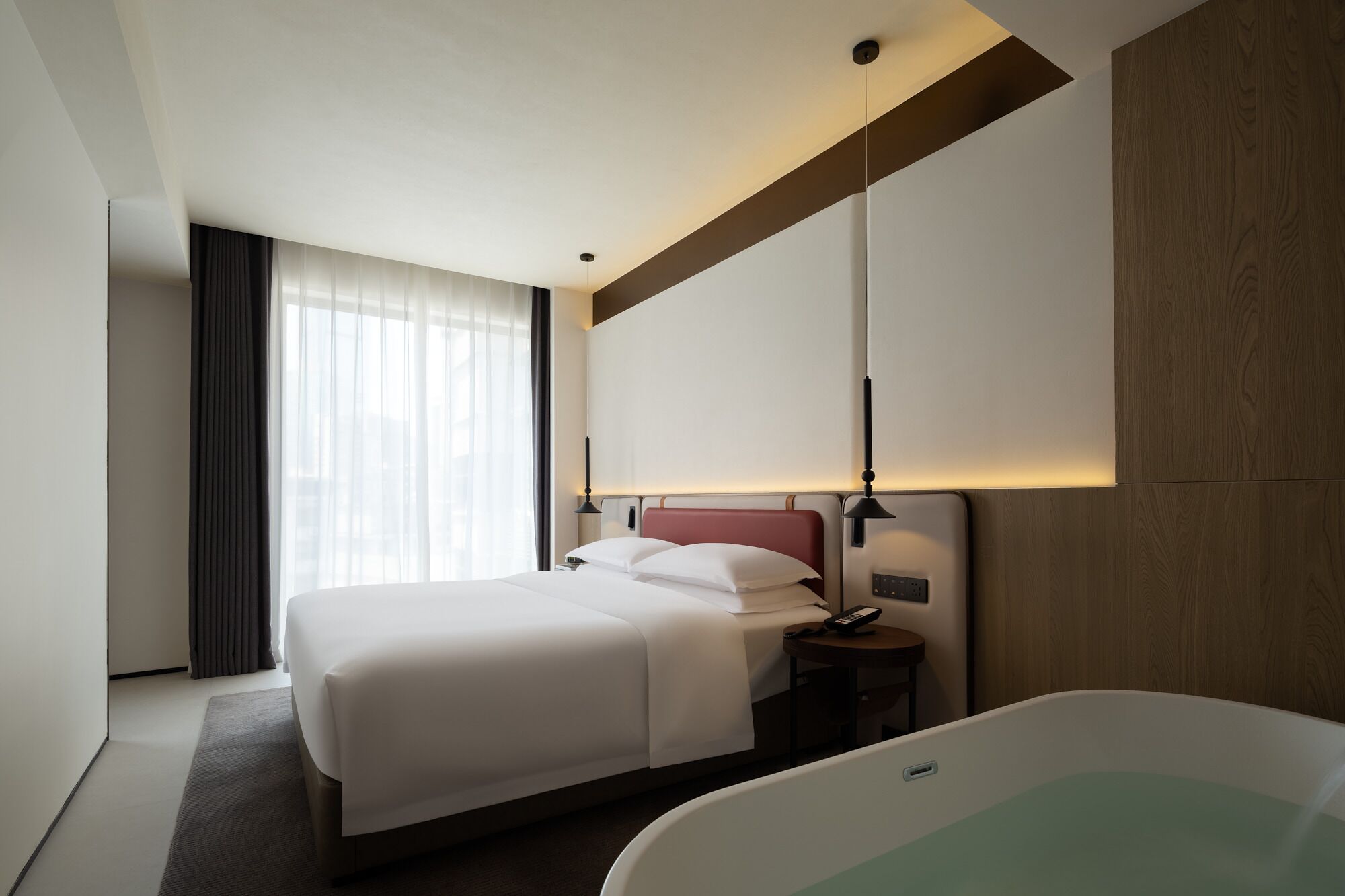 Een hotelkamer met een groot bed, een nachtkastje met een lamp, een groot raam met gordijnen en een bad.