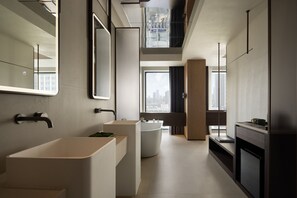 A modern bathroom with a large sink, a bathtub, and a mirror. There are windows providing natural light and a view of the city. - Book'in Hotel (Changsha)