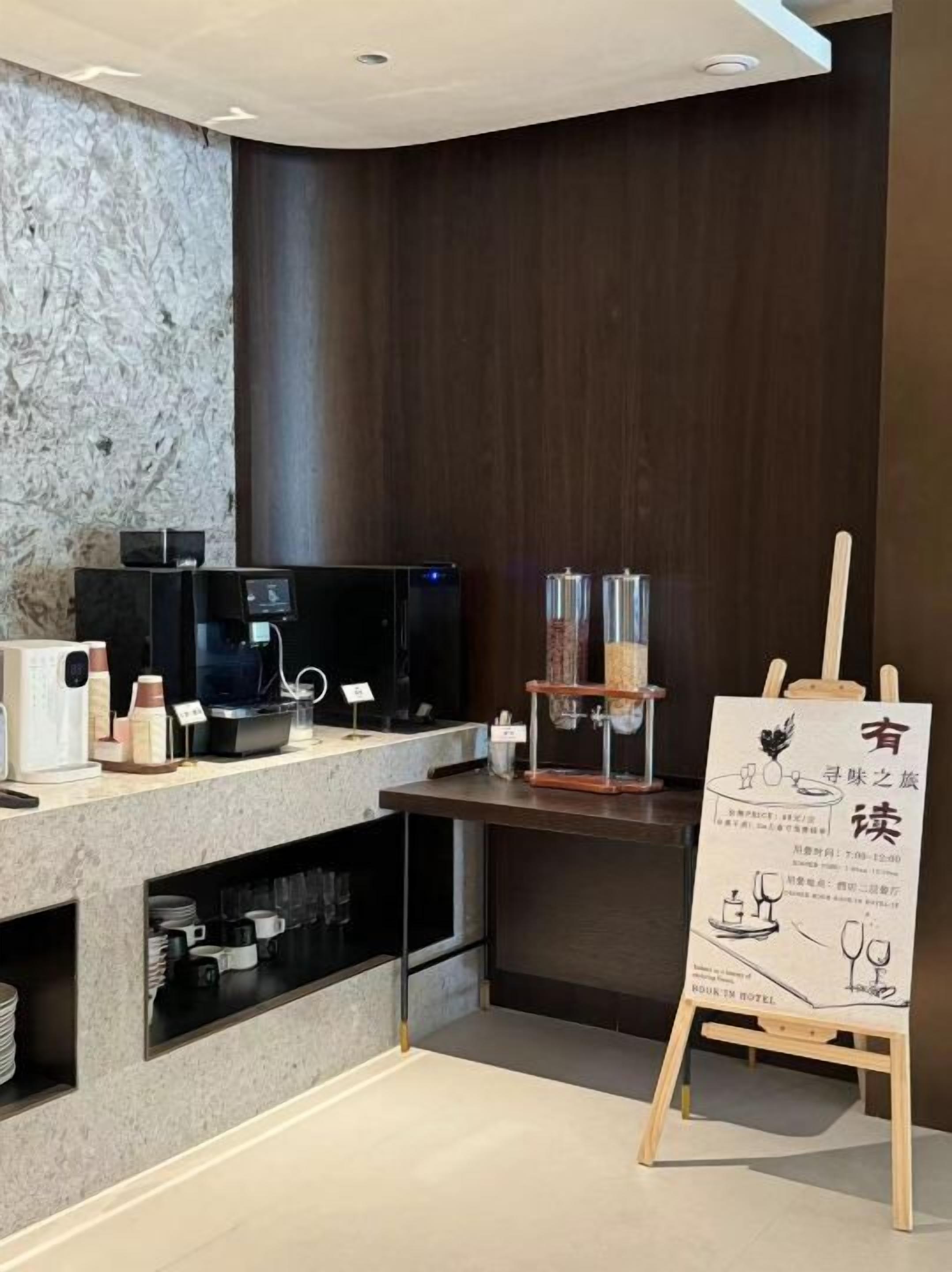 A modern hotel lobby with a marble wall, a wooden reception desk, a coffee machine, and a menu stand.