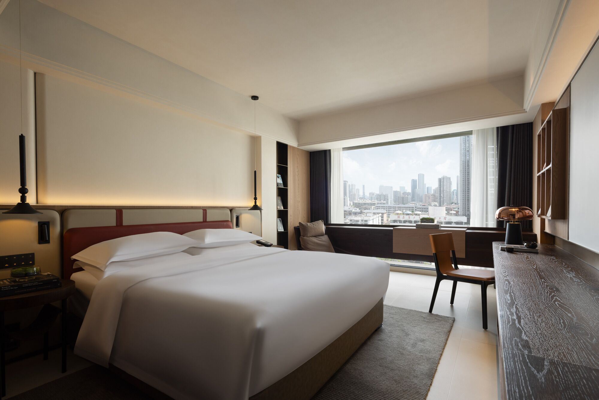 A hotel room with a large bed, a desk with a chair, a bookshelf, and a city view through the window.