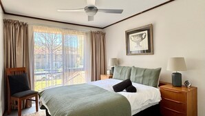 A hotel room with a bed, a chair, a nightstand, a lamp, a framed picture, and a view of the outdoors. - Little Gem in the City (Braddon)