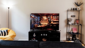 Een woonkamer met een grote televisie, een bank, een koffiezetapparaat en een plank met potten.