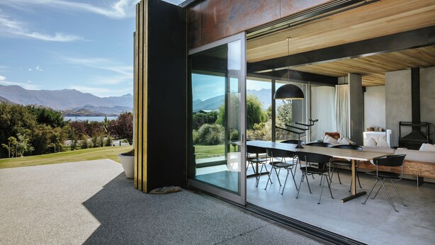 Et moderne hus med en stor glasdør, der fører til et udendørs spiseområde med bord og stole.