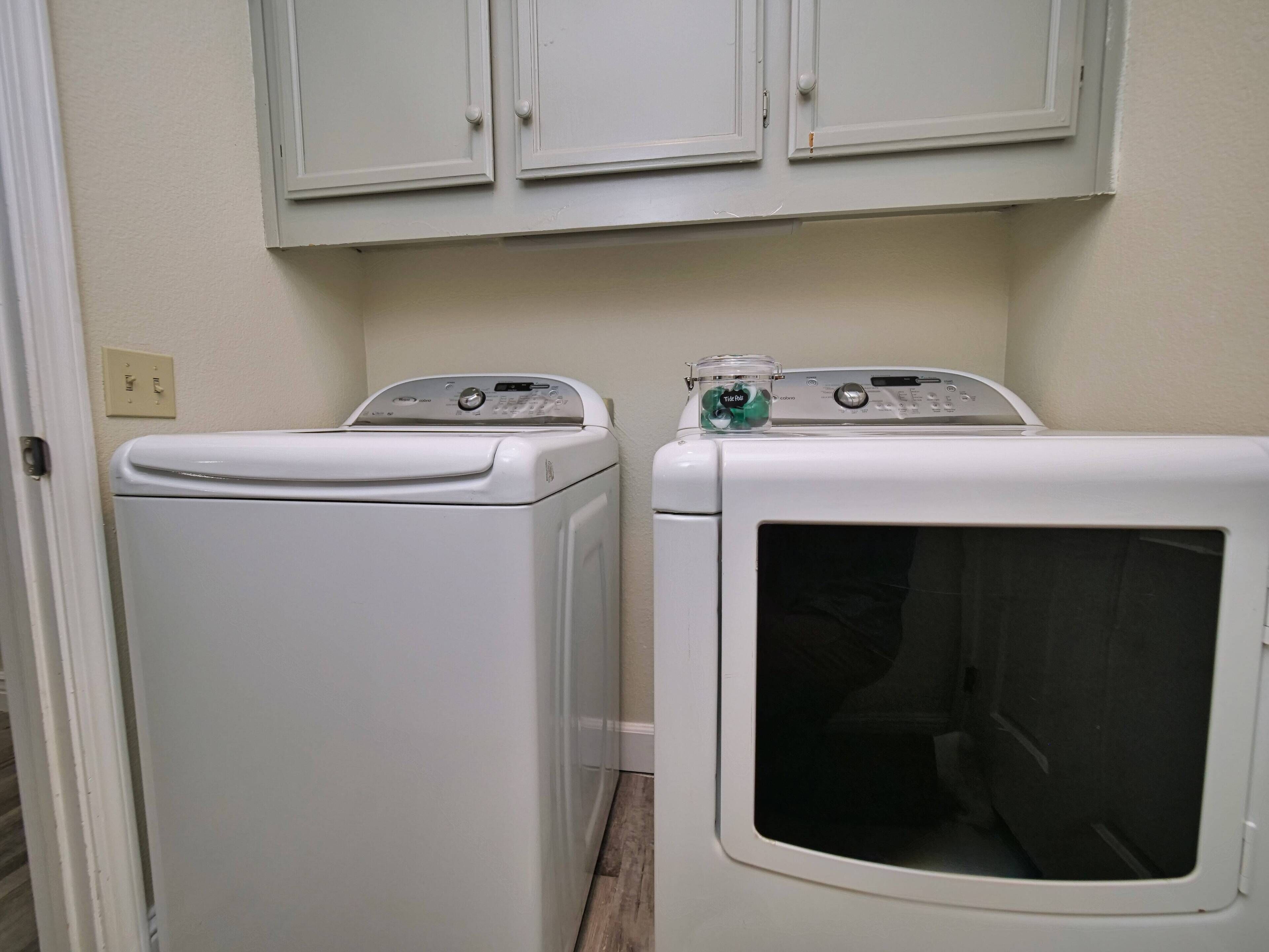 A laundry area with two white washing machines and a dryer, a white cabinet above, and a light switch on the wall.