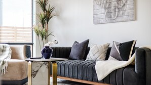 A modern living room with a dark grey sofa, a small side table, a potted plant, and a large wall art piece.