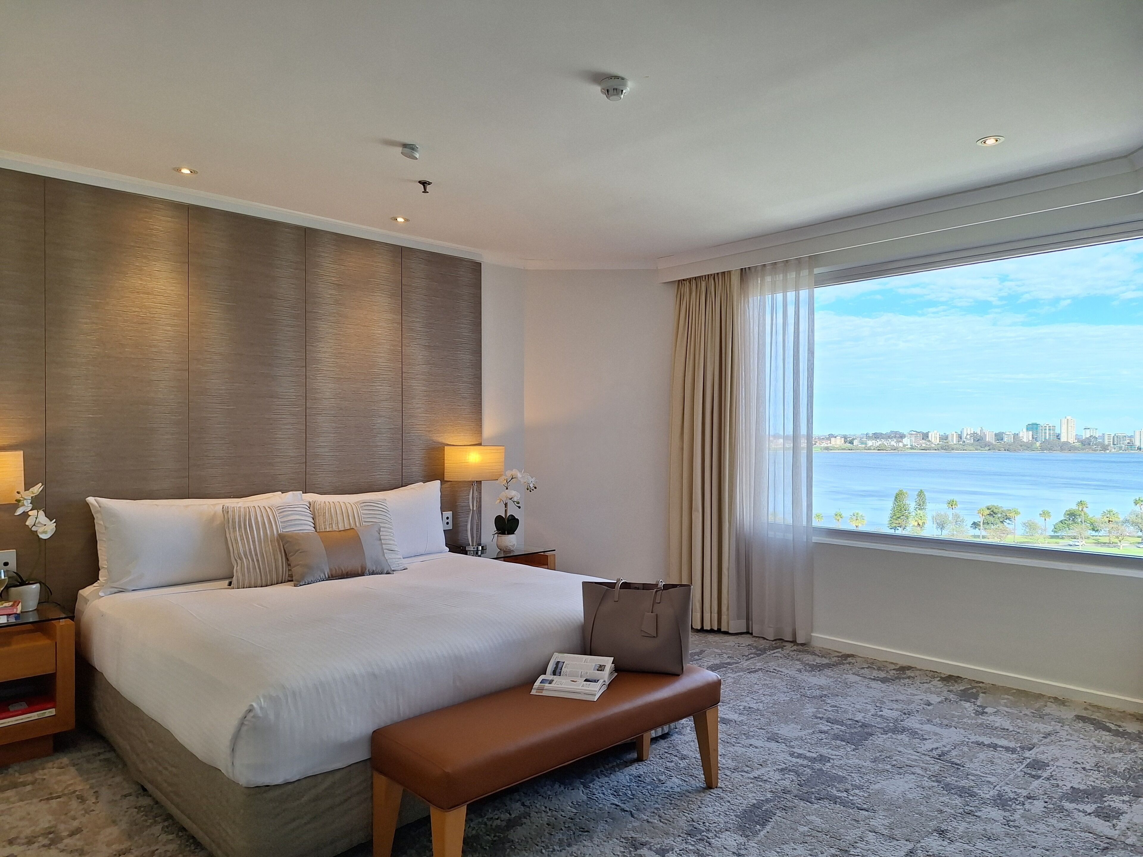 A hotel room with a large bed, bedside lamps, a bench, a window with a city view, and a ceiling with recessed lighting.