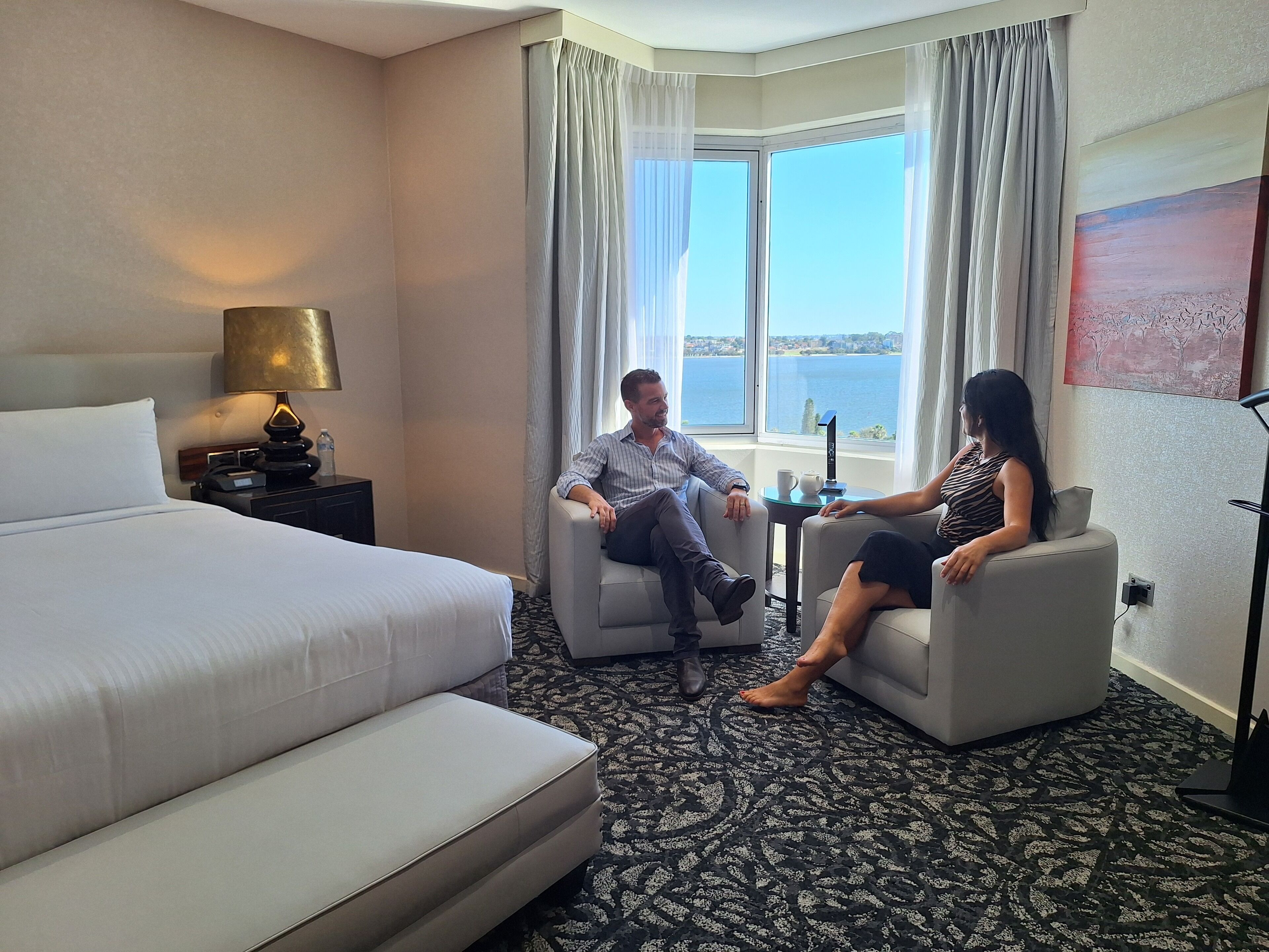 A hotel room with a large bed, two armchairs, a small table, and a view of the sea through a large window.