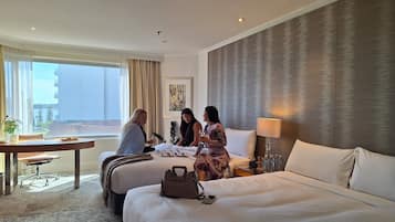 A hotel room with a large bed, a desk with a chair, a window with curtains, and a view of buildings outside.