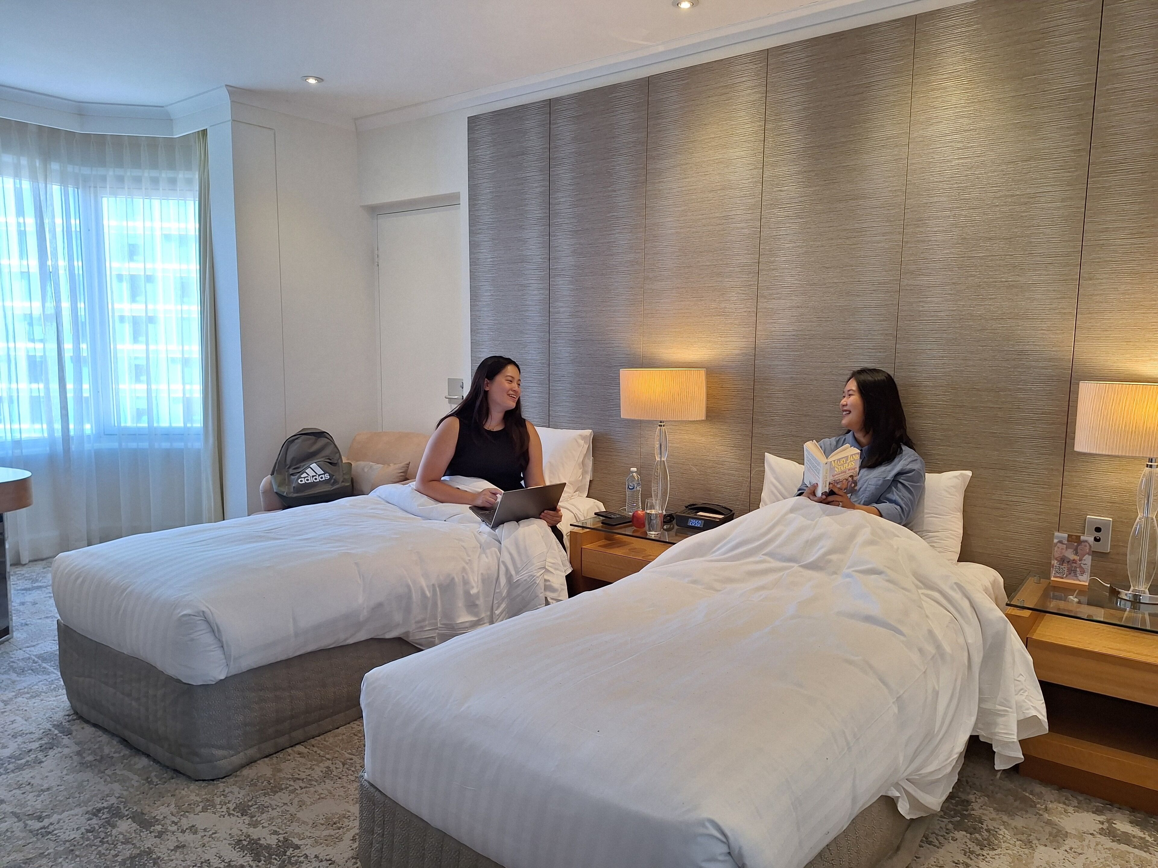 Ein Hotelzimmer mit zwei Betten, einem Nachttisch mit Lampe, einer Wasserflasche und einem Koffer.