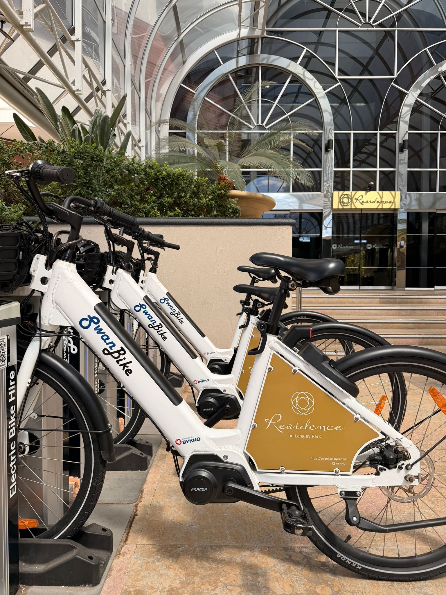 Bicicletas eléctricas con la marca 'Swan Bike' y 'Electric Bike Hire' en un lugar con un cartel que indica 'Residence', señalando un servicio de alquiler.