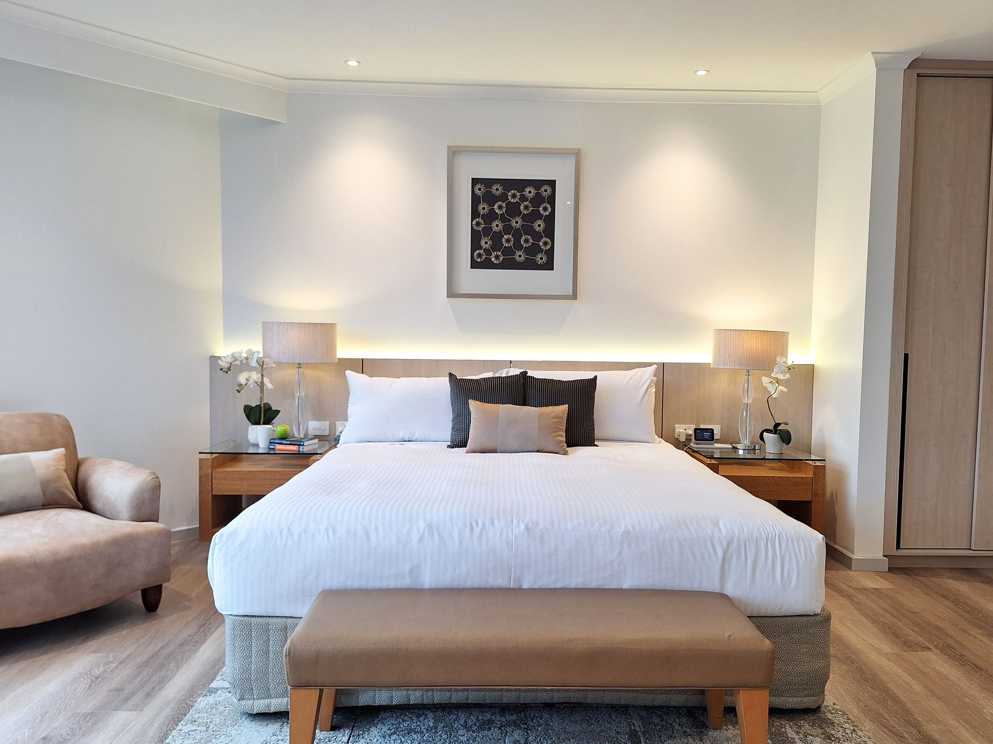A modern hotel room with a large bed, two bedside tables with lamps, a chair, and a wooden bench.
