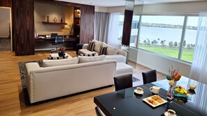 A modern living room with a sofa, coffee table, and dining table with pastries. A large window offers a view of a body of water and greenery. - Residence on Langley Park (East Perth)