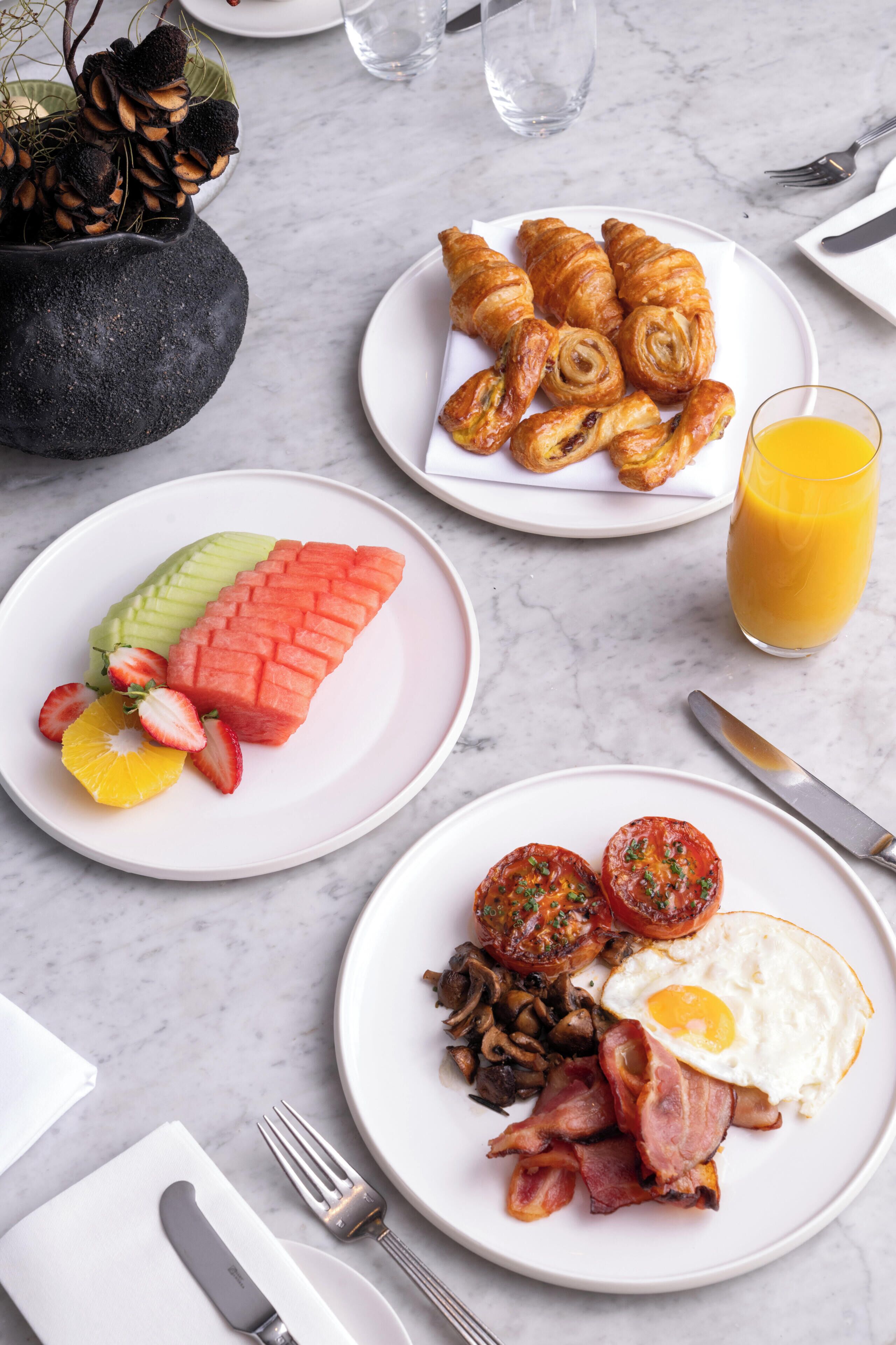 Une table dressée pour le petit-déjeuner avec des croissants, des tranches de pastèque, des fraises, des tranches d’orange et un verre de jus d’orange.