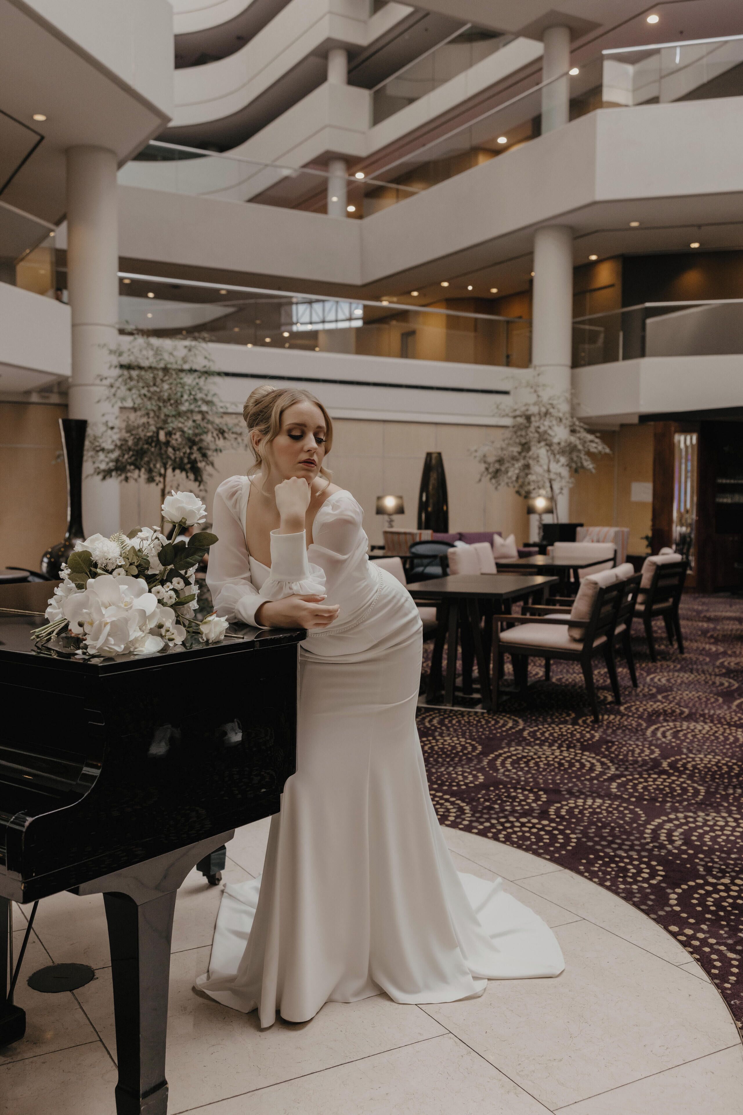 Une femme vêtue d’une robe blanche se tient à côté d’un grand piano dans le hall spacieux et à plusieurs niveaux d’un hôtel à l’architecture moderne, dont le sol est recouvert d’une moquette.