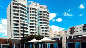 A swimming pool with clear blue water, surrounded by lounge chairs and umbrellas, with modern buildings in the background. - Residence on Langley Park (East Perth)