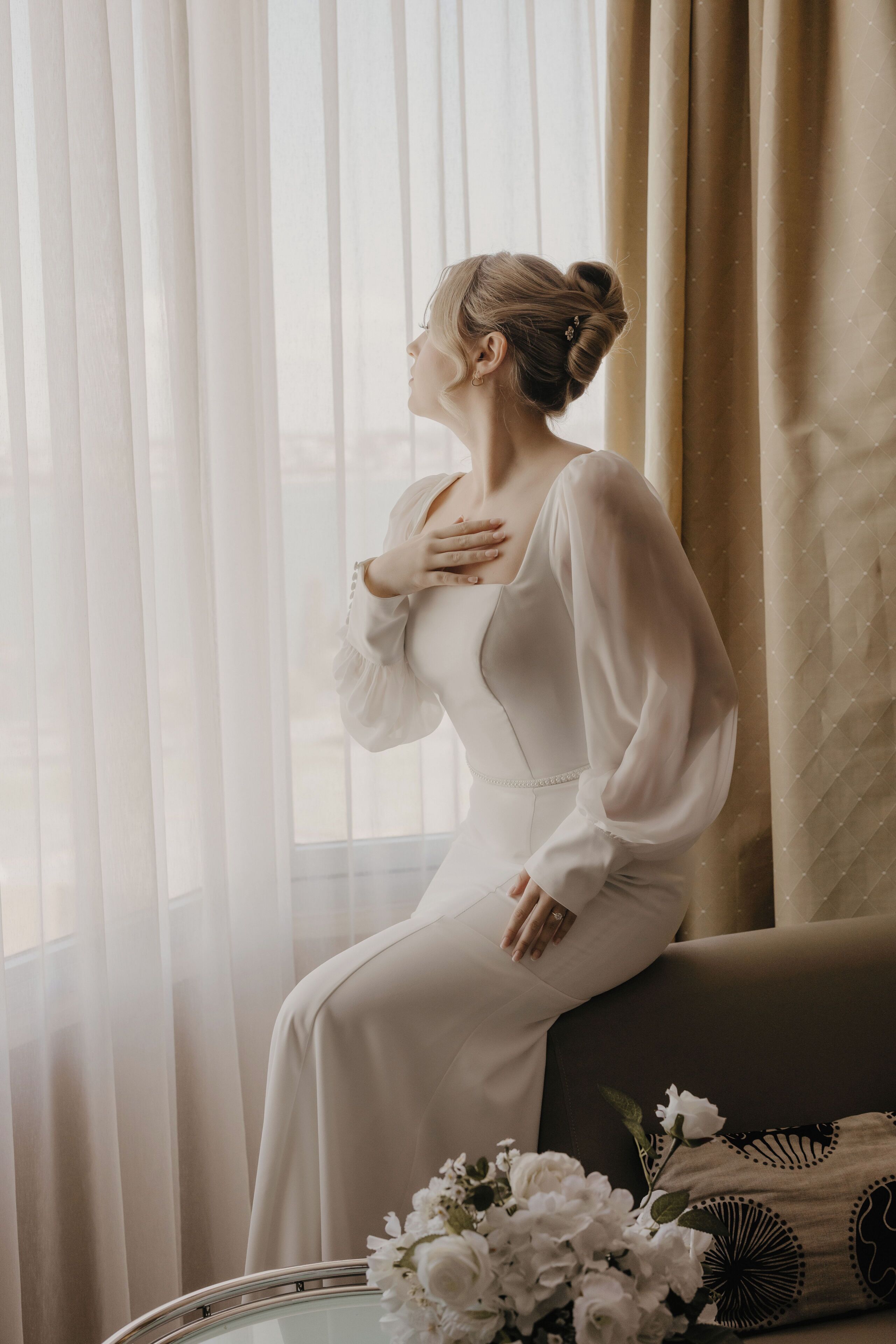 A person seated on a sofa in a room with sheer curtains and a floral arrangement on a table.