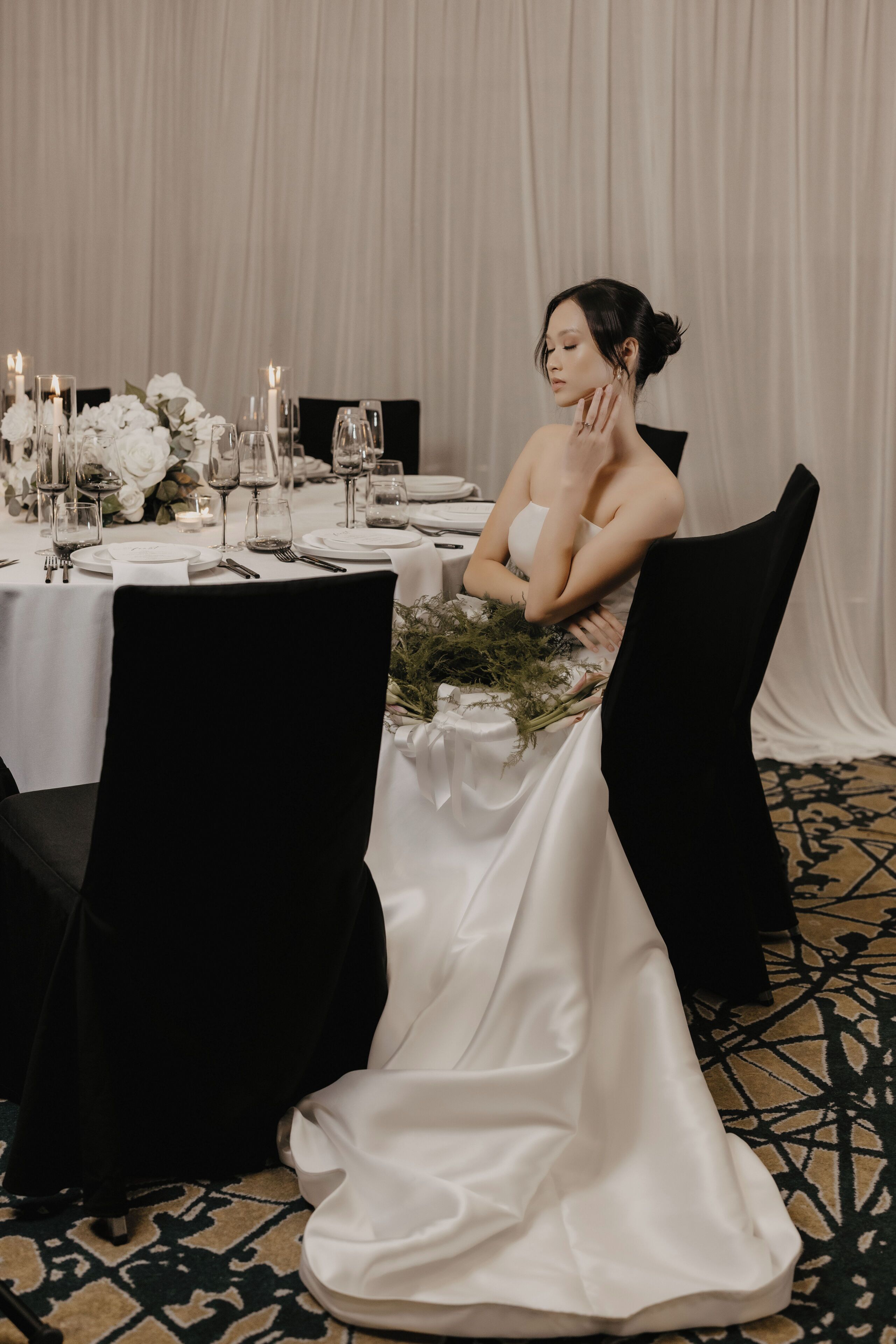 Una persona sentada a una mesa con mantel blanco, rodeada de sillas negras, en una habitación con alfombra estampada y un fondo de cortina blanca.