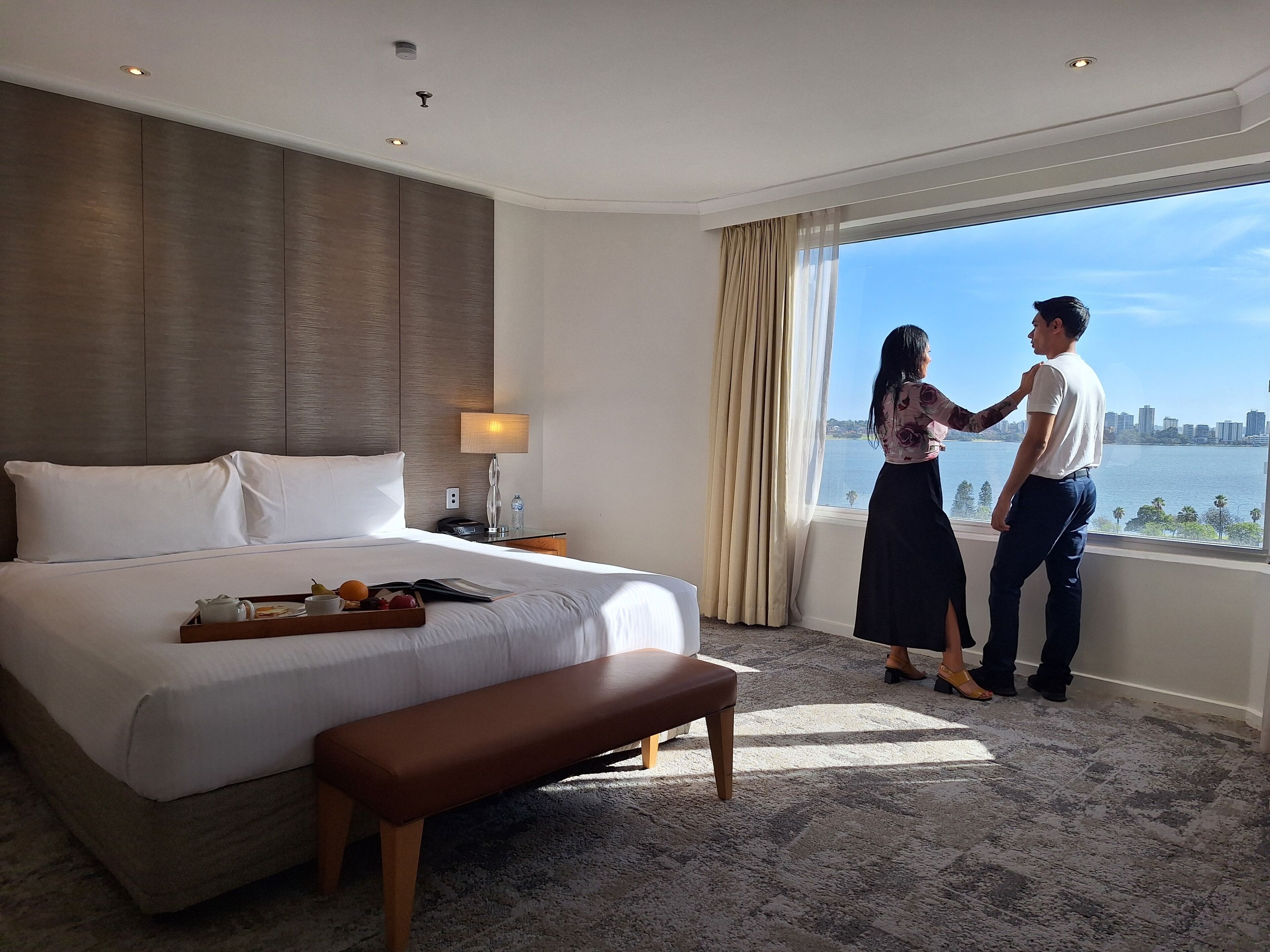 A hotel room with a large bed, a bedside table with a lamp, a tray with fruits and a book, and a view of a cityscape through the window.