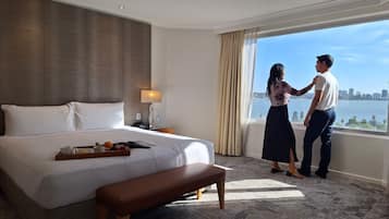 A hotel room with a large bed, a bedside table with a lamp, a tray with fruits and a book, and a view of a cityscape through the window.