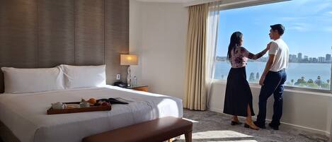 A hotel room with a large bed, a bedside table with a lamp, a tray with fruits and a book, and a view of a cityscape through the window.