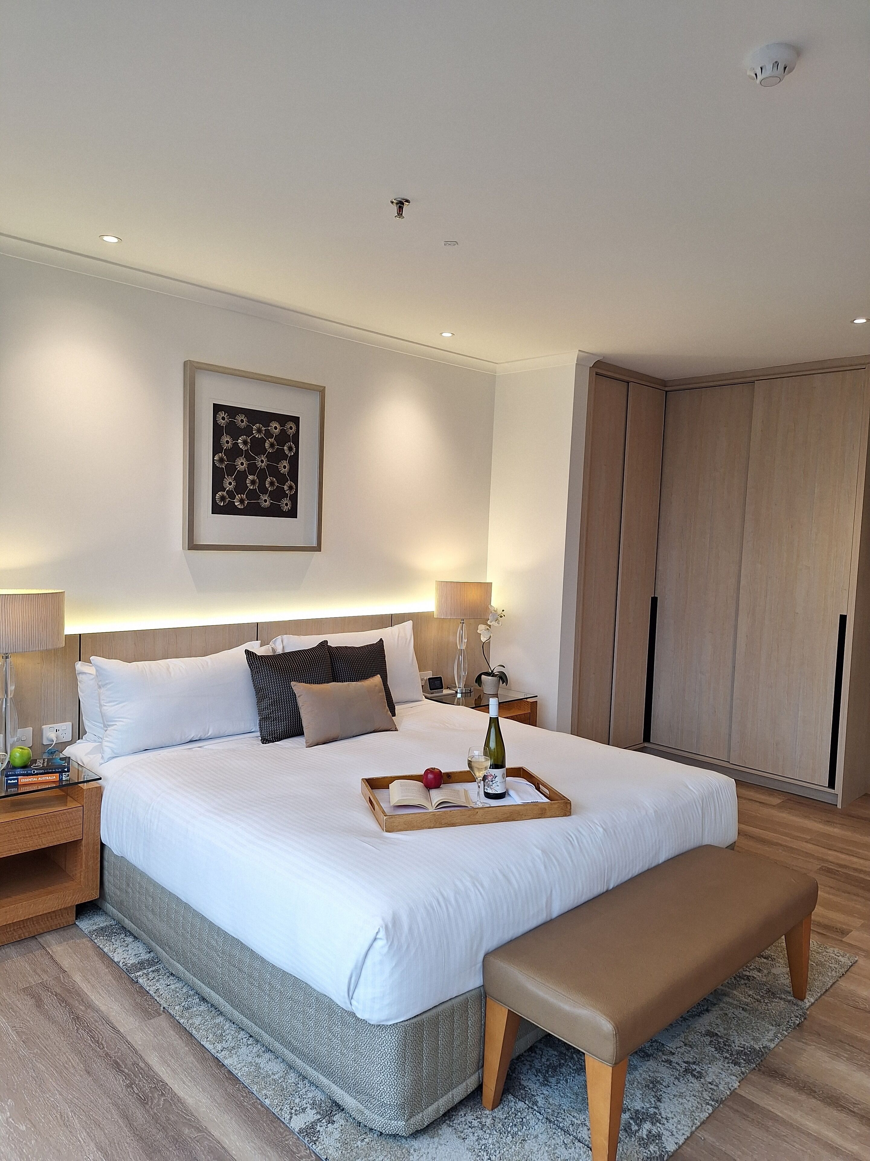 A modern hotel room with a large bed, bedside tables, a bench, a wardrobe, and a framed artwork on the wall.