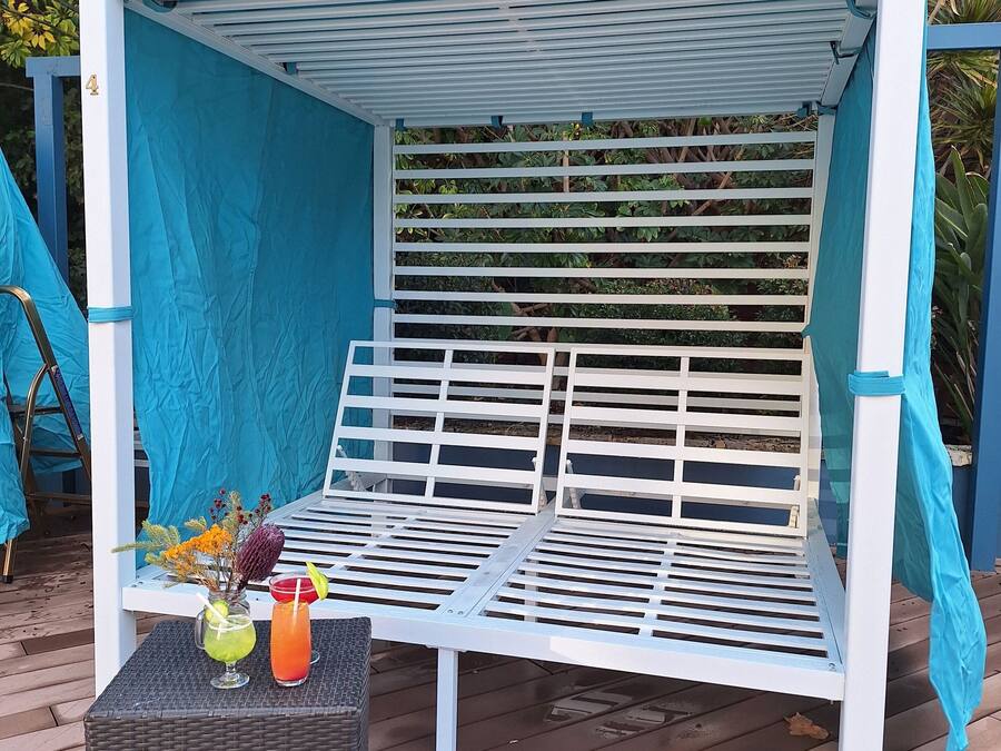 A white outdoor seating area with a blue canopy, a wicker table with drinks, and a backdrop of greenery.