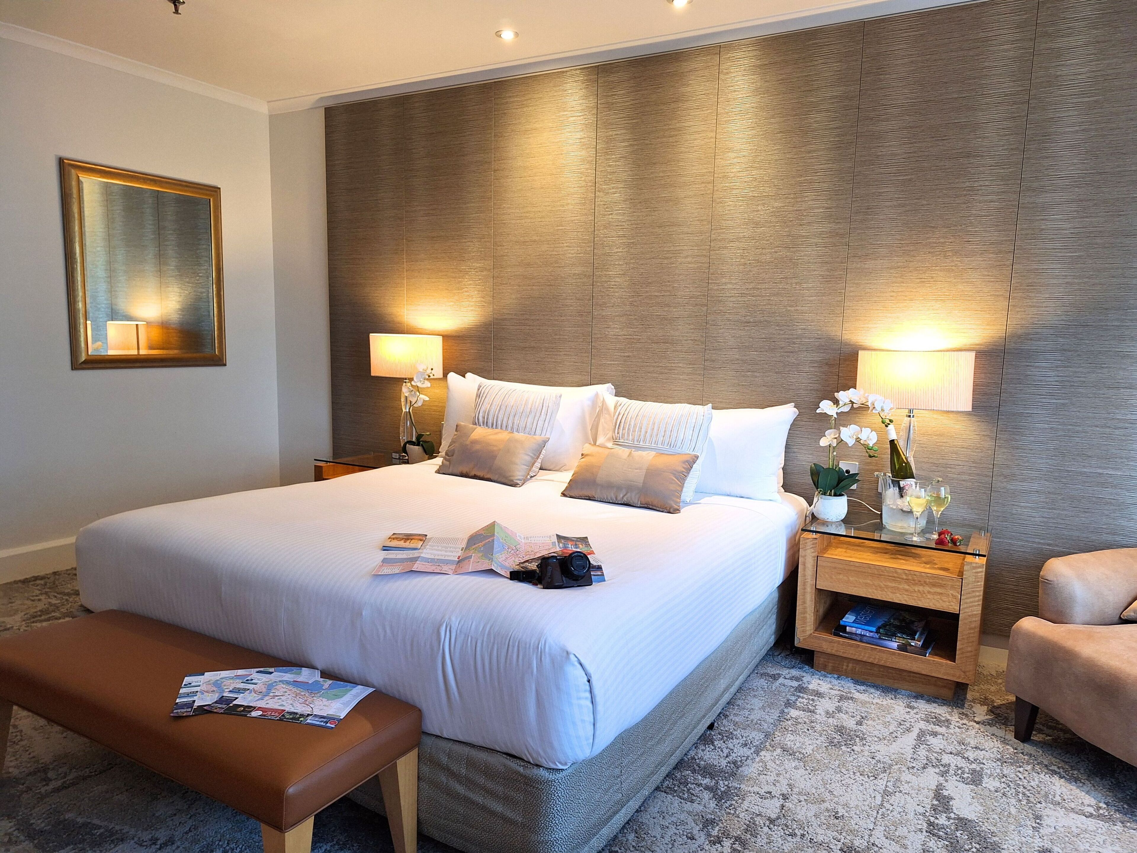 A modern hotel room with a large bed, two bedside tables with lamps, a bench, and a small side table with a bottle and glasses.