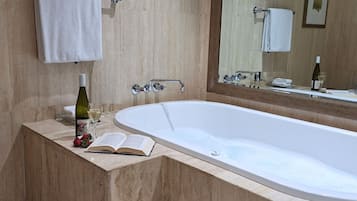 A modern bathroom with a large bathtub, a vanity with a wine bottle and book, and a mirror reflecting a towel and a framed picture.