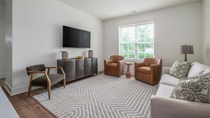 Uma sala de estar com sofá, duas poltronas, mesa de centro, TV na parede, janela com persianas e tapete estampado.