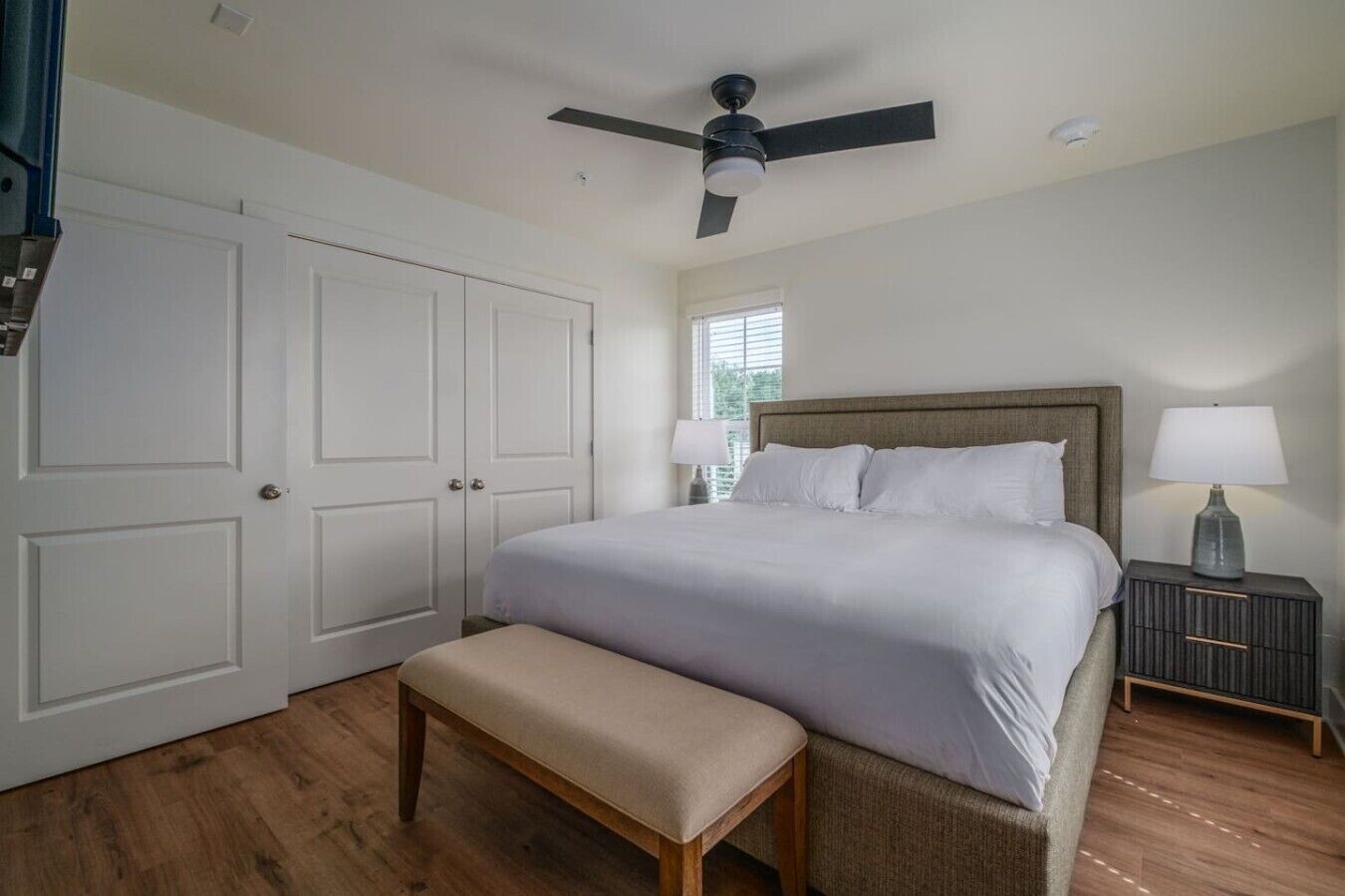 A bedroom with a bed, a bench, a nightstand, a lamp, a ceiling fan, and a window.