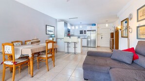A modern living room with a grey sofa, wooden dining table, and chairs. A kitchen with white cabinets and a stainless steel refrigerator is visible in the background. - Apartment Navio by Interhome (Aguadulce)