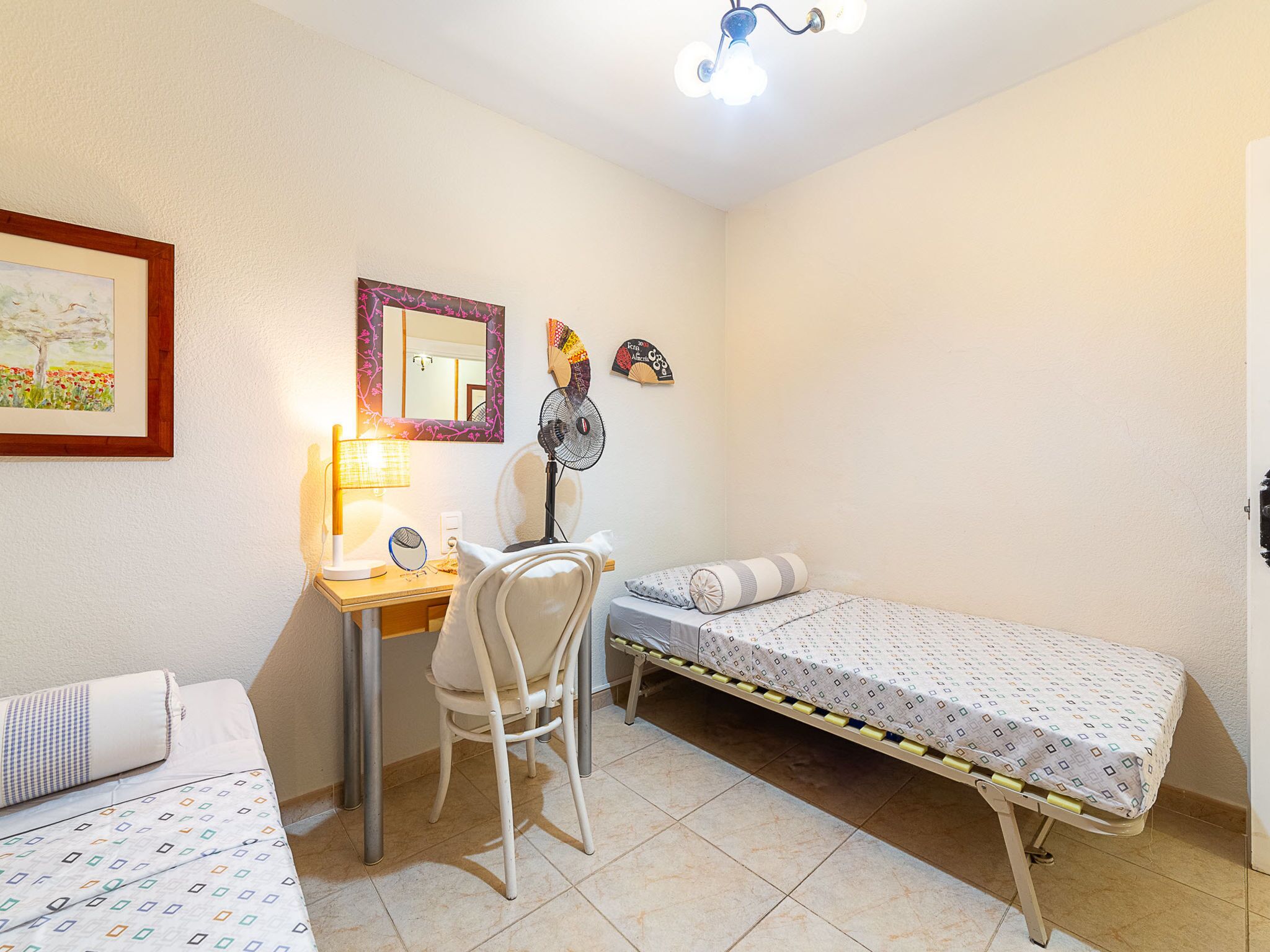 Una habitación pequeña y luminosa con escritorio, silla, dos camas, espejo y ventilador de techo.